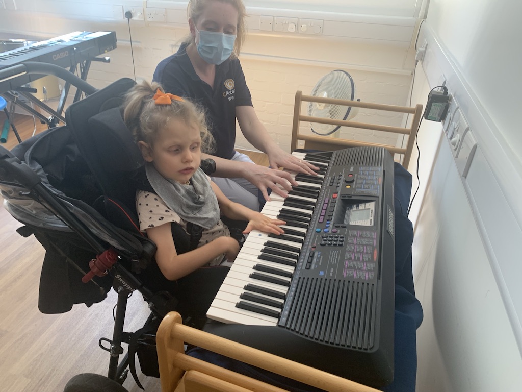 Young girl with a disability sitting in a push chair playing a musical keyboard with the aid of a therapist sitting next to her 