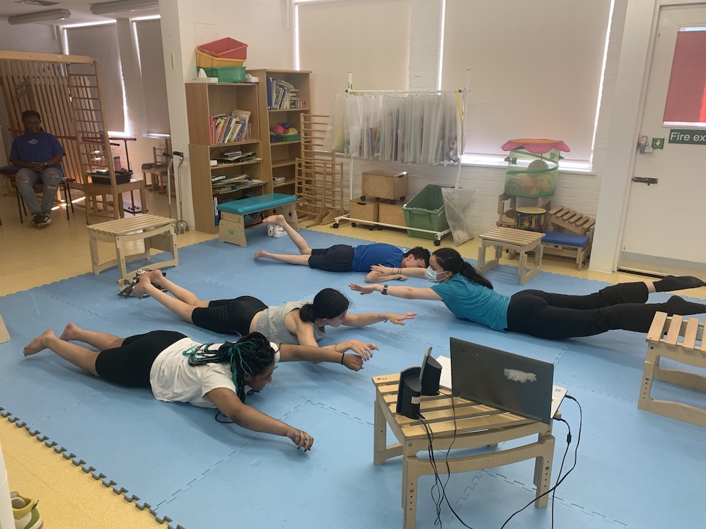 Three students lying on their stomachs being instructed by a therapist to lift their legs and arms in the air