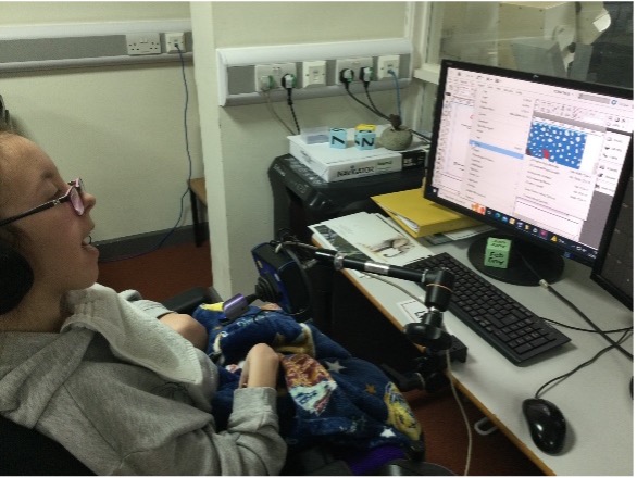 Libby sitting in her wheelchair in front of a computer using her bespoke computer mouse / joystick to create print designs for a customer.  

