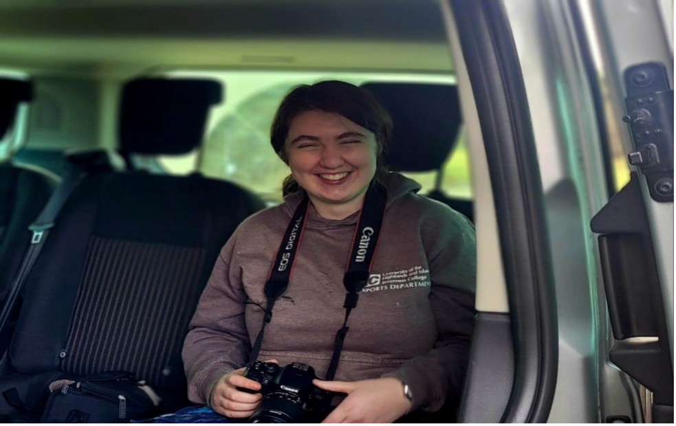 Photo of a lady with a broad smile sitting in a car, het camera around her neck 