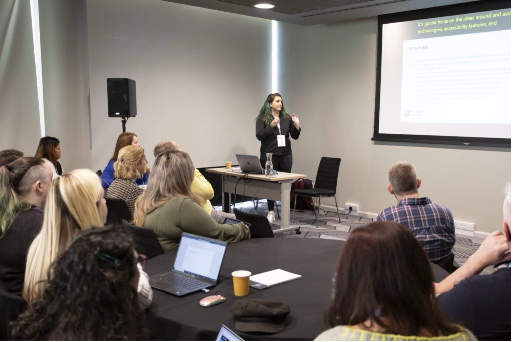 Presenter in front of screen, talking to room of delegates who are sitting round tables
