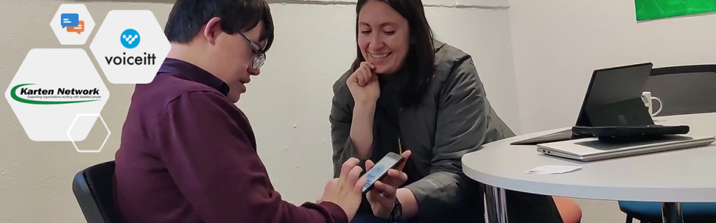 A Nuvoic project participant sitting, holding a mobile phone in his hand. A project team member sitting beside him, explaining how to use the Voiceitt app 