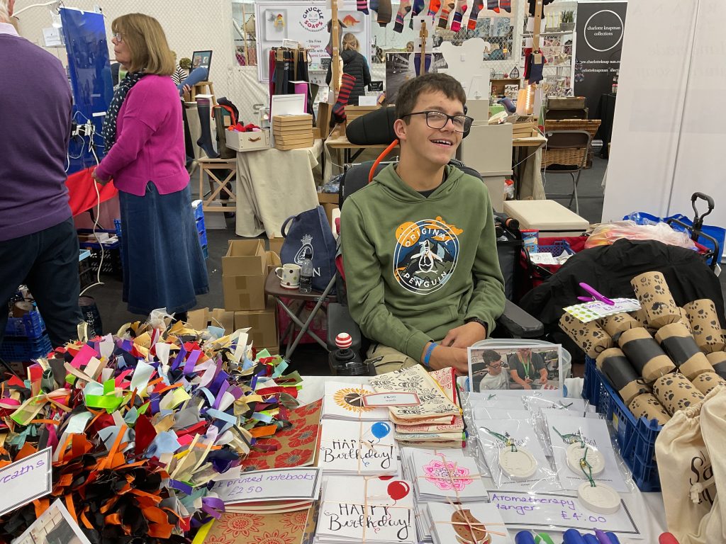 Ben, a young man sitting at the Treloar stall at at Dummer Fair. On the table in front of him are birthday cards, crackers and many other items for sale. 