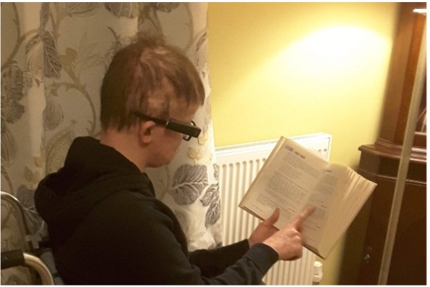 A young man, sitting reading a book. He is wearing the OrCam MyEye device on his glasses