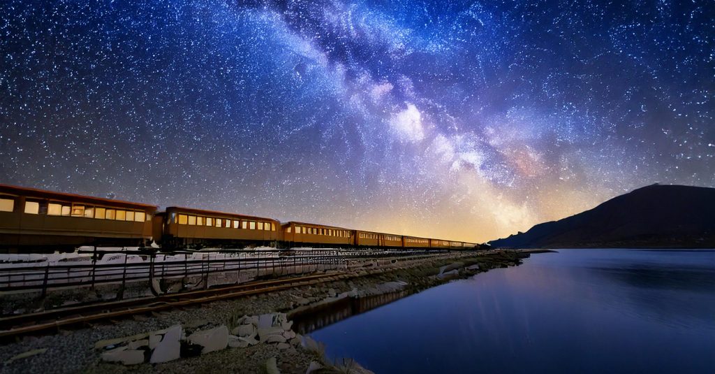 A picture of a train travelling next to a lake at night, with the milky way shining above.
