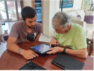 An elderly woman and a young man sit at a wooden table, interacting with a tablet. The woman points at the screen while the man attentively assists her. A second tablet and case are on the table, with a living room and a cat in the background.