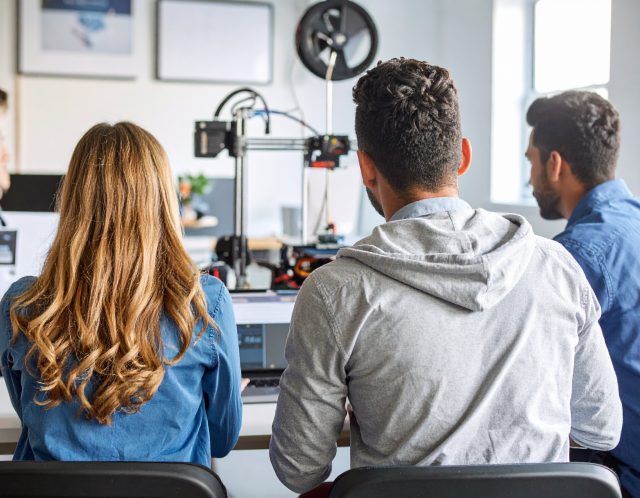 Three students sit at a desk, viewed from behind, working together in a classroom setting. In front of them is a 3D printer in operation, with laptops open. The room is bright, with natural light coming through the windows, and posters hang on the wall in the background.