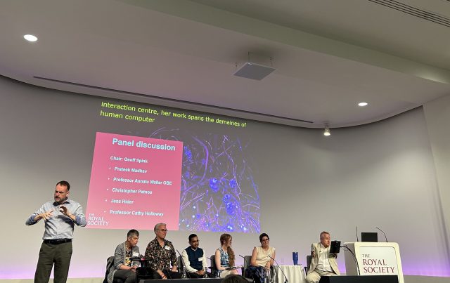 A panel discussion at The Royal Society on Disability Technology, with six speakers seated on stage beneath a projected slide listing their names. A British Sign Language interpreter stands to the left, mid-gesture. The screen behind them displays the event branding and a visual of neural-like digital art. Live captions are visible above the slide.
