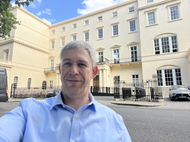Martin Pistorius in a light blue shirt takes a selfie outside a grand cream-colored building with tall windows and black railings. The sky is blue with scattered clouds. The setting is The Royal Society in London.