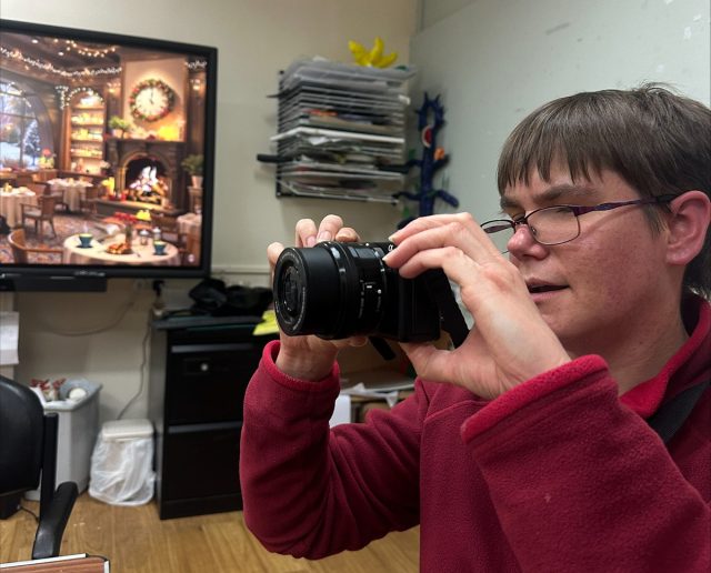 A person wearing glasses and a red fleece holds a digital camera up to their eye, preparing to take a photo. They are indoors, with a cozy, festive scene of a decorated dining room displayed on a large screen in the background. Art supplies and shelves can be seen in the surrounding room.