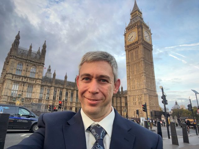 Martin Pistorius, smiling in a suit and tie, sitting in his wheelchair in front of Big Ben and the Palace of Westminster in London on a cloudy day.