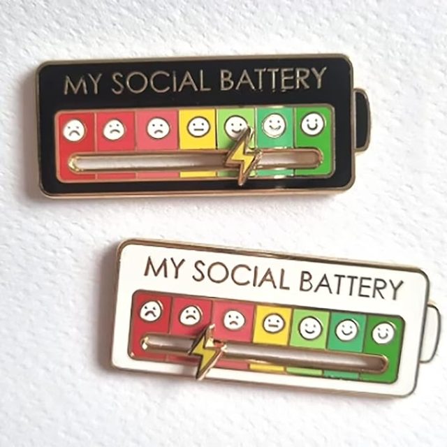 A wearable slider badge labeled “My Social Battery,” featuring a visual scale of five emoji faces from sad to happy, with a lightning bolt slider currently pointing to the green (fully charged) end of the scale.