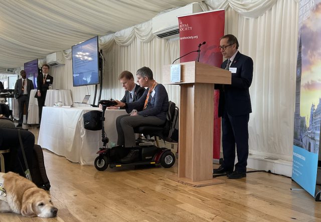 Professor Sir Bernard Silverman FRS speaking at the launch of The Royal Society’s Disability Technology report at the House of Commons. He stands at a lectern beside a Royal Society banner, while panellists, including a wheelchair user, are seated behind him. A guide dog rests on the floor in the foreground.