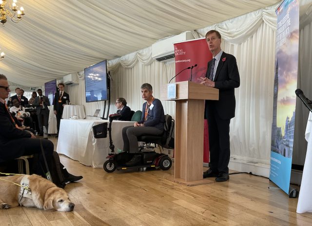Sir Stephen Timms MP, Minister of State for Social Security and Disability, speaking at the launch of The Royal Society’s Disability Technology report at the House of Commons. He stands at a lectern beside a Royal Society banner, with panellists seated behind him and a guide dog resting on the floor in the foreground.