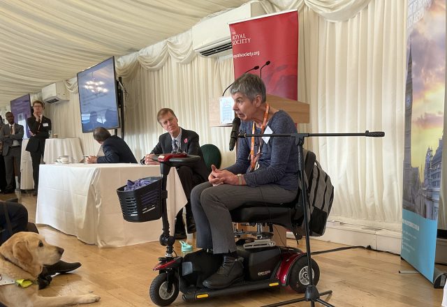 Professor Annalu Waller OBE FRSE speaking at the launch of The Royal Society’s Disability Technology report at the House of Commons. She is seated in a power wheelchair with a microphone, addressing attendees beside a Royal Society banner. A guide dog rests on the floor in the foreground.