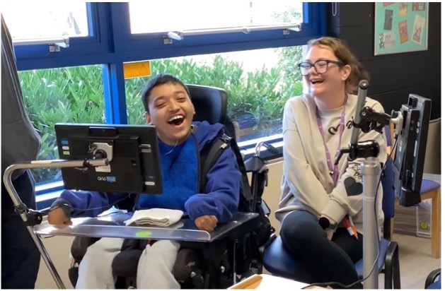 A student using a communication device smiles and laughs alongside a staff member.