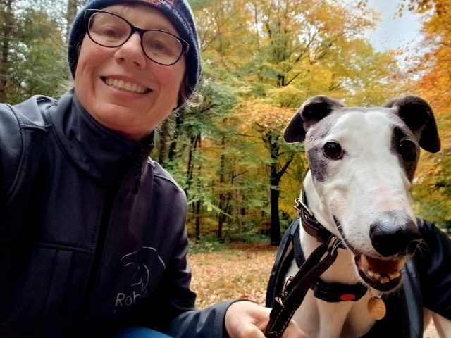 Dawn Green smiling outdoors with her dog, a white and grey greyhound named Cuthbert. She is wearing glasses, a blue hat, and a dark jacket. Autumn trees with yellow and orange leaves fill the background.