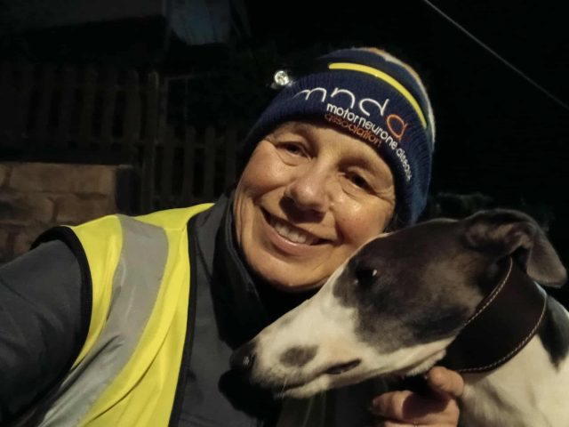 Dawn Green smiling at night while holding her greyhound, Cuthbert. She is wearing a Motor Neurone Disease Association hat and a reflective yellow vest. The photo is warmly lit against a dark background.